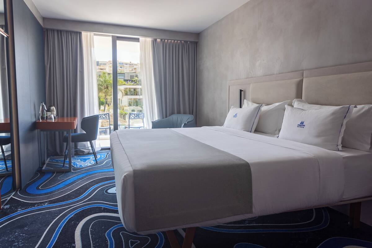 Ein Hotelzimmer mit einem großen Bett, einem Schreibtisch und einem Stuhl. Durch das Fenster ist ein Blick nach draußen.