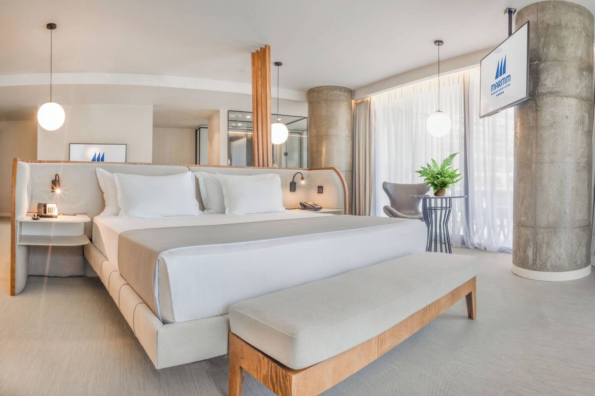 Ein modernes Hotelzimmer mit einem großen Bett, Nachttischen, einer Bank, einer Topfpflanze und einem großen Fenster mit Vorhängen.