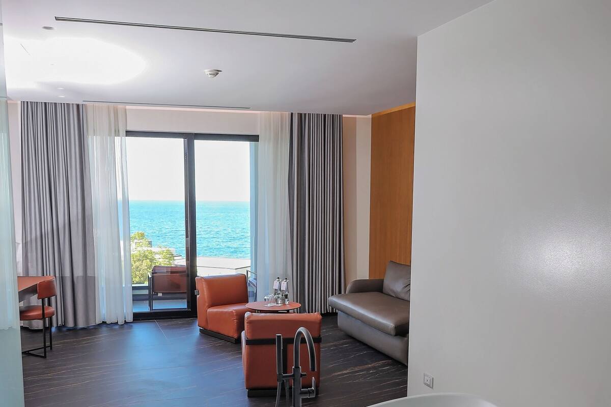 Ein modernes Hotelzimmer mit einer großen Badewanne, einer Sitzecke inklusive Sofa und Sesseln, sowie einem Balkon mit Meerblick.