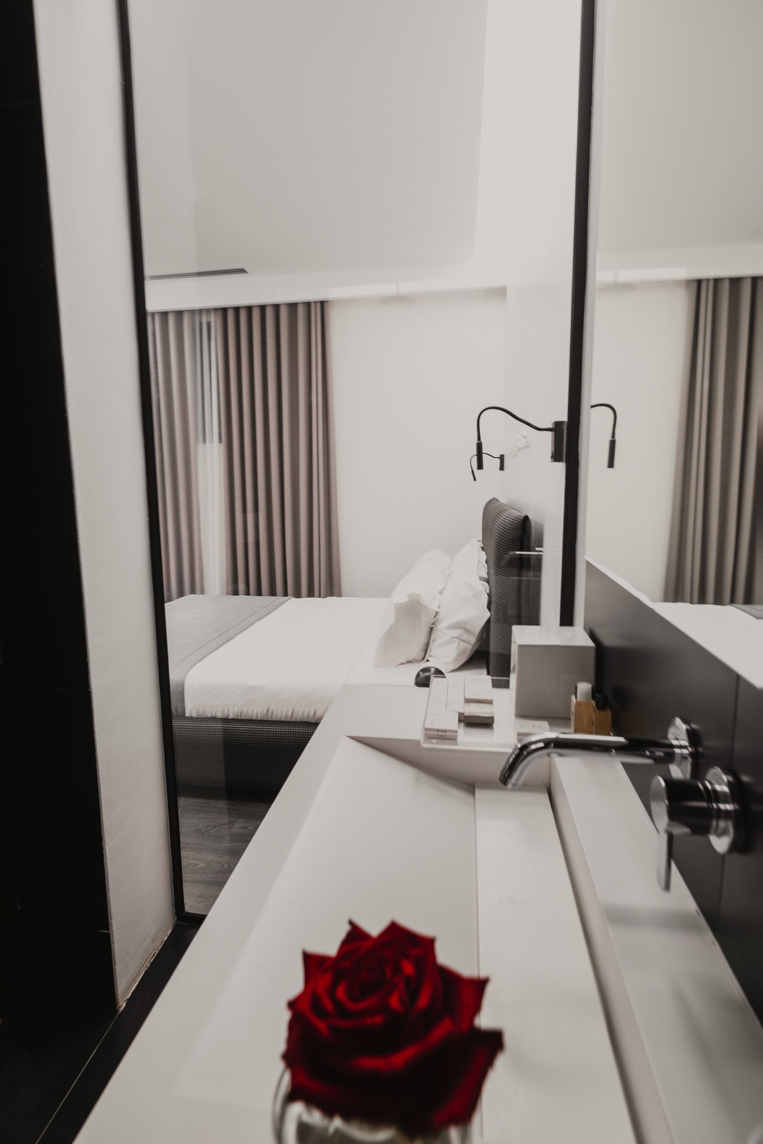 A modern hotel room with a large mirror, a white bed, a bedside table with a bottle, and a red rose on the counter.
