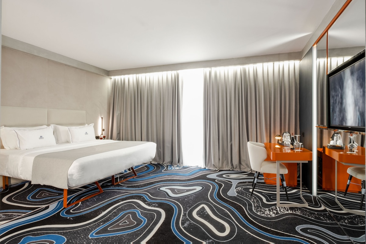 A hotel room with a bed, a desk, a chair, a television, and a large window with curtains.