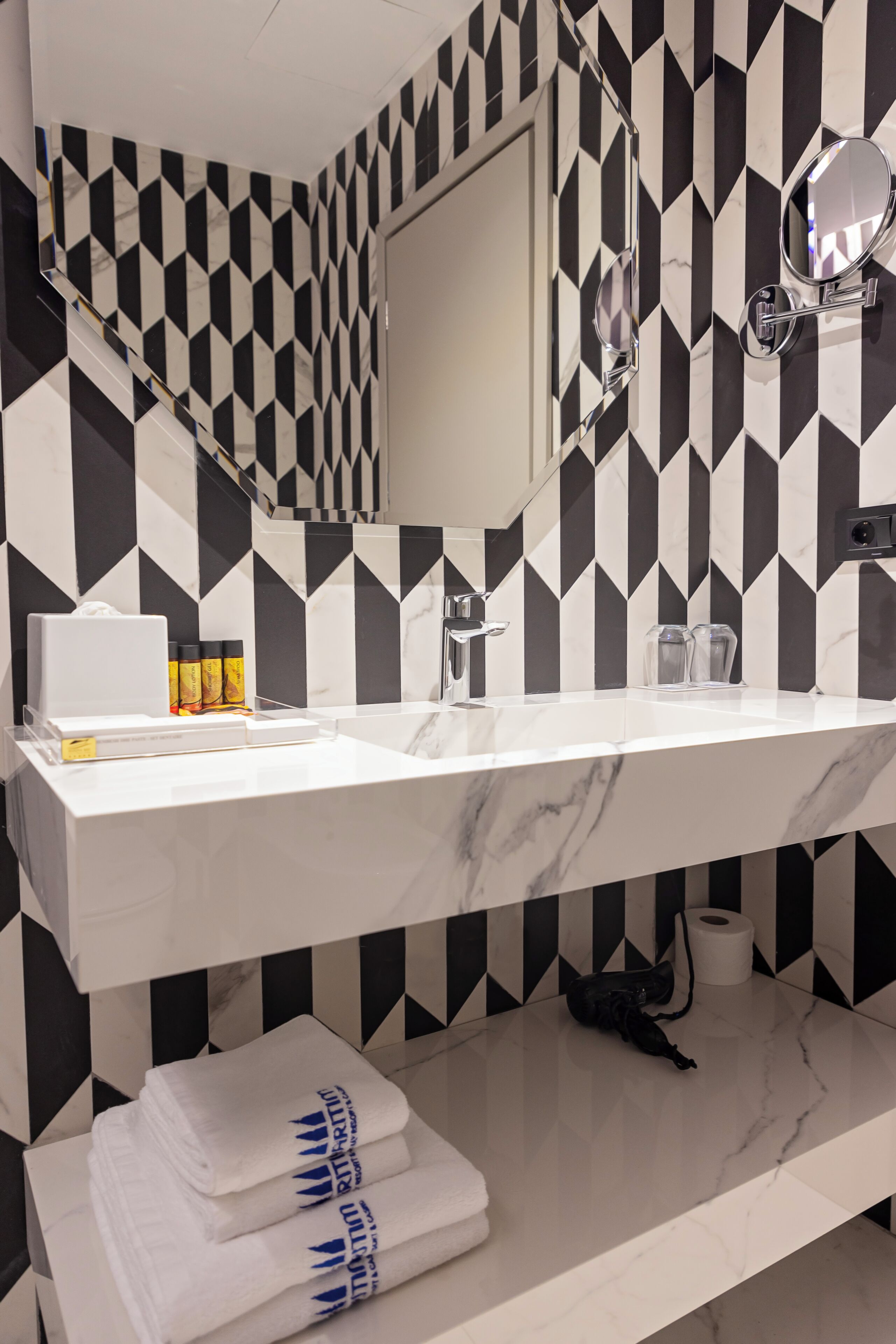 A bathroom with a black and white geometric tile backsplash, a white marble sink, a mirror, and neatly folded towels.
