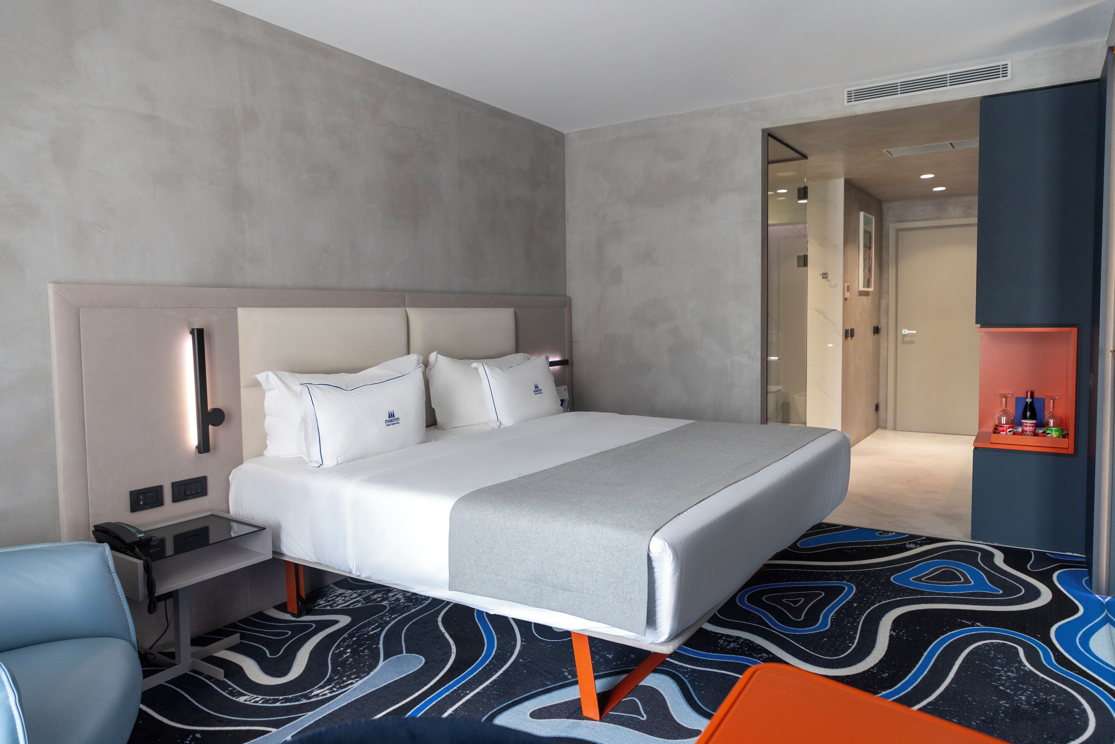 A modern hotel room with a large bed, a bedside table with a phone, a chair, and a carpet with a geometric pattern.