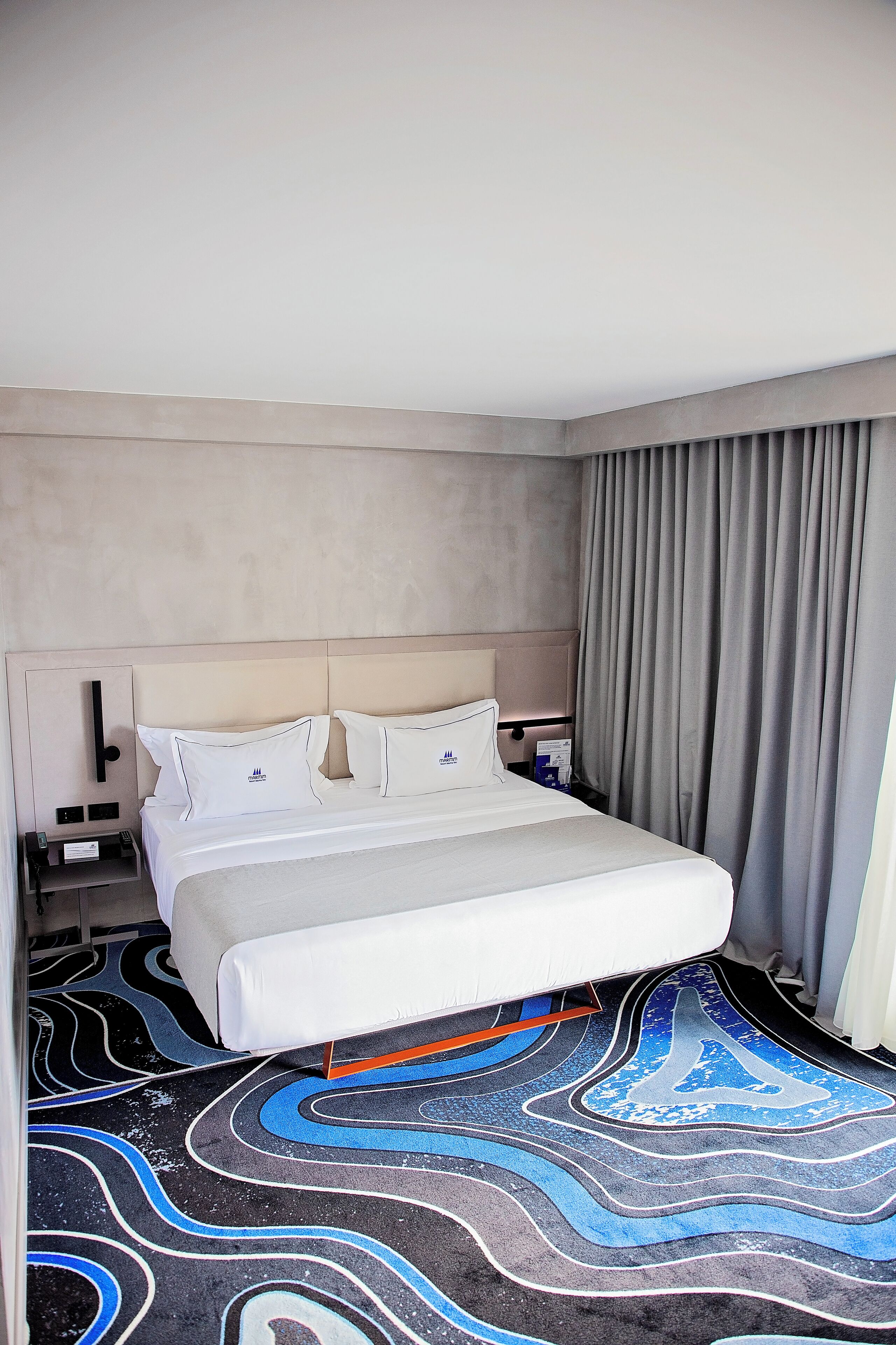 Habitación de hotel con cama, mesita de noche, lámpara, ventana con cortinas y alfombra estampada.