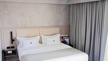 Habitación de hotel con cama, mesita de noche, lámpara, ventana con cortinas y alfombra estampada.