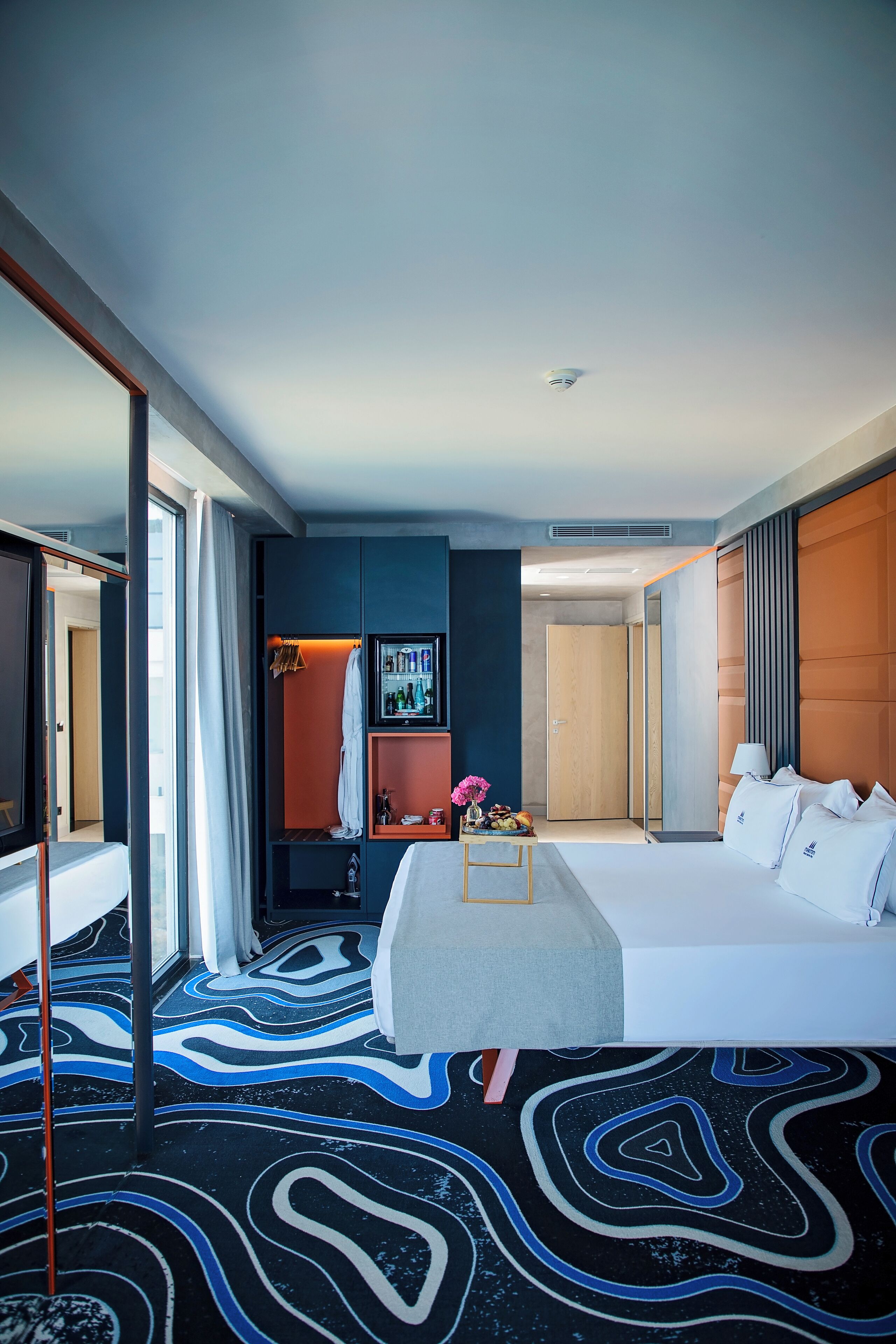 Een moderne hotelkamer met een groot bed, een kledingkast, een kaptafel met spiegel en een zichtbare badkamer door een open deur.