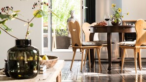 A dining area with a wooden table and chairs, a vase with flowers, and a view of greenery outside.