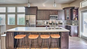 A modern kitchen with dark wood cabinets, a large island with stools, stainless steel appliances, and a view through large windows. - Lavish 3 BR La Quinta Oasis < 2 Mi to Old Town (La Quinta)