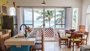 Une cuisine équipée d’un évier et d’un réfrigérateur, un coin repas avec des chaises et une table, et une vue sur l’océan grâce à de grandes fenêtres.