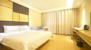 A hotel room with a large bed, a desk with a chair, a small table with a lamp, and a curtain-covered window.
