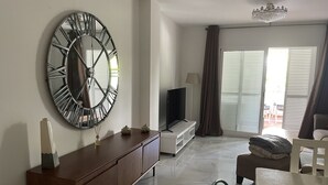 A modern living room with a large wall clock, a flat-screen TV, and a sliding glass door leading to an outdoor area. - Live in Paradise (Marbella)