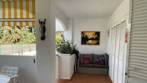 A balcony with a white chair, a sofa with colorful cushions, a painting, and a view of trees and a fence. - Live in Paradise (Marbella)