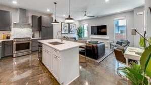 A modern kitchen with a central island, stainless steel appliances, pendant lighting, and a dining area with a sofa and table. - New, Luxury Home| Private Roof Deck (Nashville)
