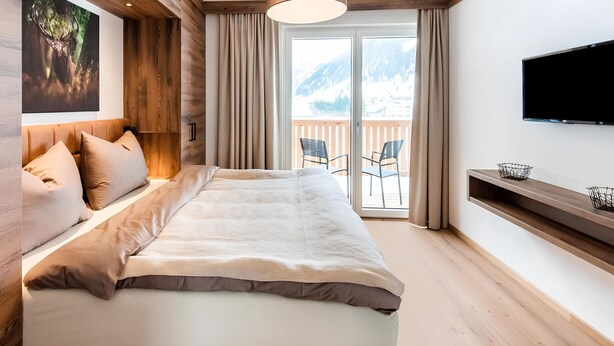 A modern bedroom with a large bed, wooden beams, a ceiling light, a TV, and a balcony with chairs.
