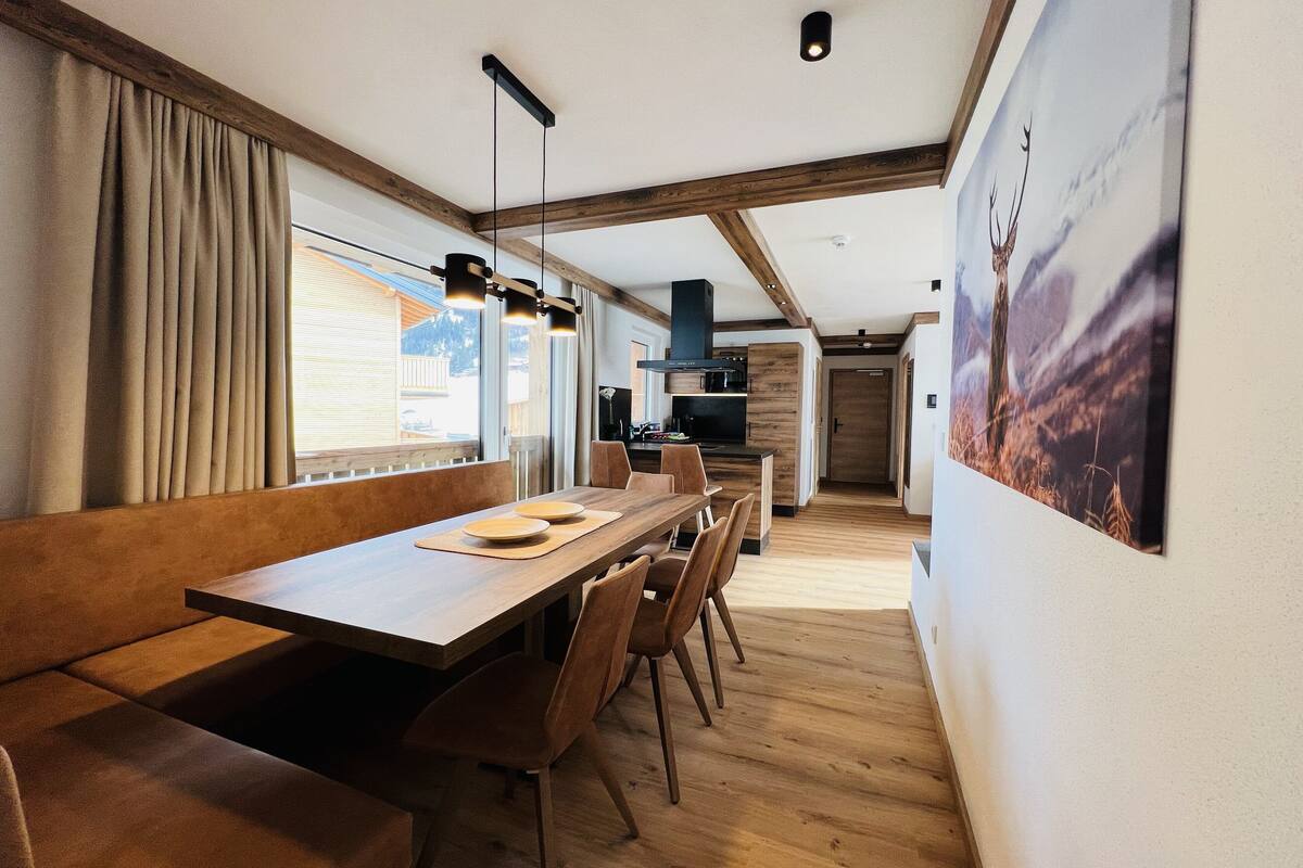 A modern dining area with a wooden table and chairs, a kitchen in the background, and a large window with curtains.
