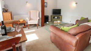 A living room with a brown leather sofa, a white armchair, a wooden dining table with a wine bottle and glass, a glass coffee table, and a television on a wooden stand. - La'l Skaithe (Kirkby Stephen)