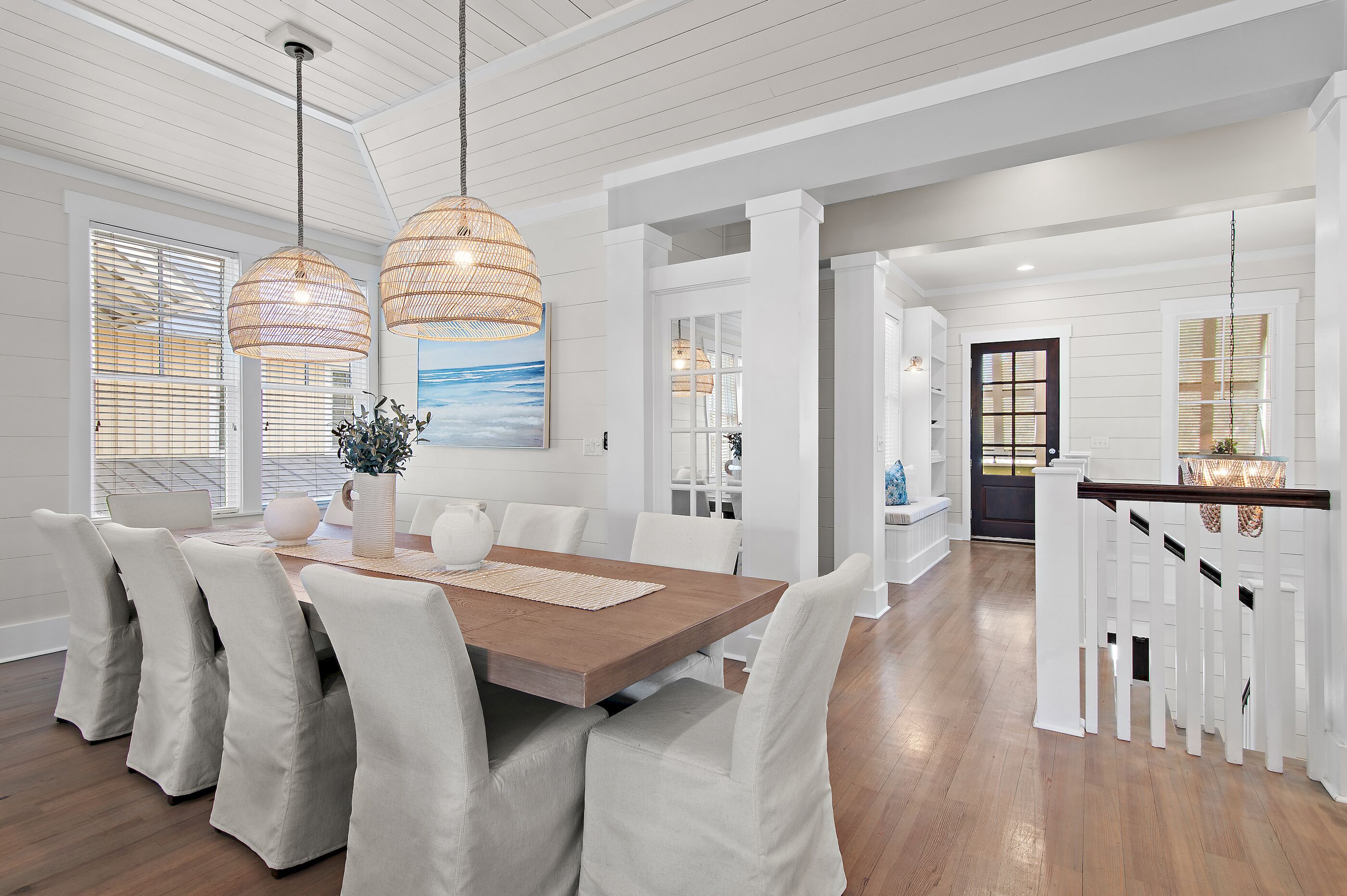 A dining area with a wooden table and white upholstered chairs, a hanging rattan light fixture, and a view of the kitchen through an open doorway.