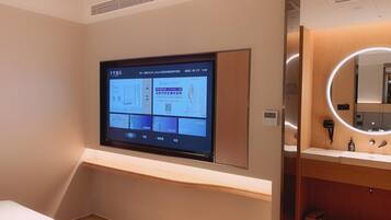 A hotel room with a mounted TV displaying a hotel website, a wooden vanity with a mirror, and a digital thermostat.