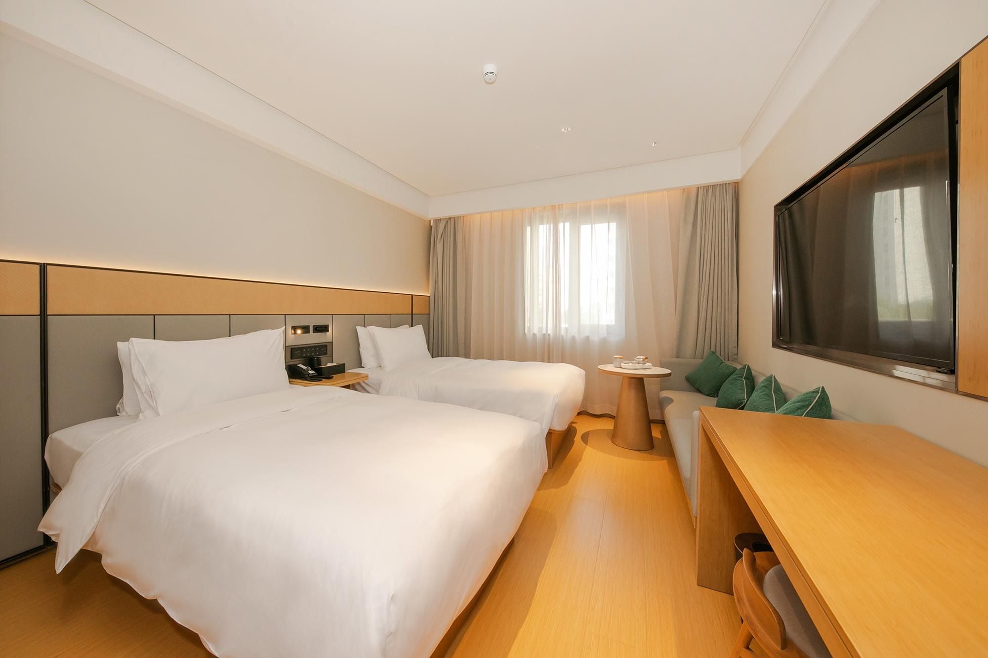 A hotel room with two beds, a desk, a small table, and a large window with curtains.