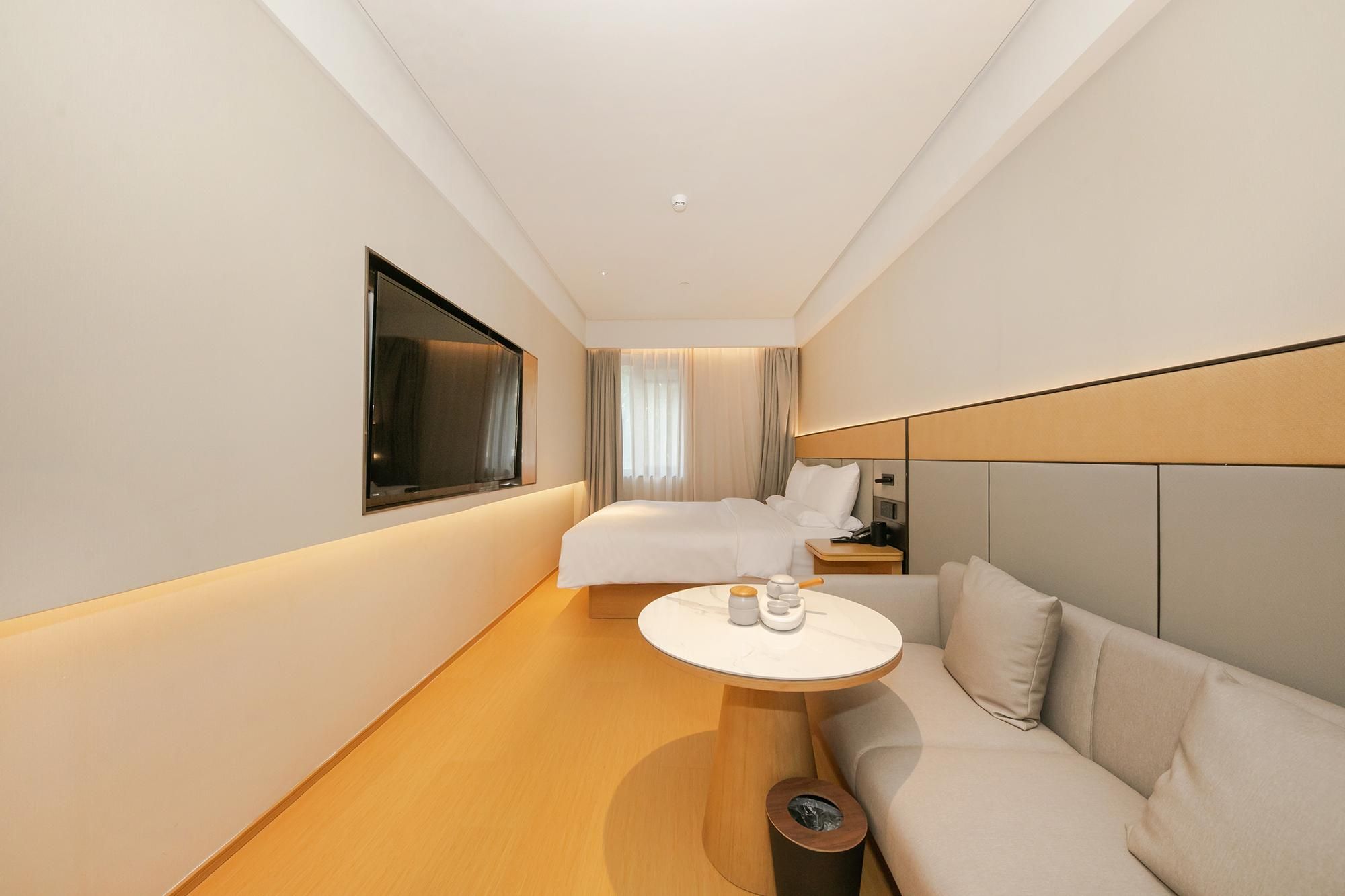 Quarto de hotel com cama, sofá, mesinha, televisão e janela com cortinas.
