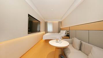 Quarto de hotel com cama, sofá, mesinha, televisão e janela com cortinas.