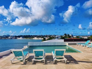 A pool area with lounge chairs and a view of the sea and cityscape.