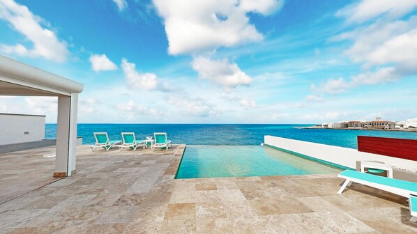 A rooftop pool with a view of the ocean, lounge chairs, and a red wall.