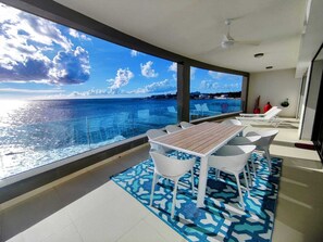 A balcony with a wooden table and white chairs, overlooking a body of water with a clear sky.