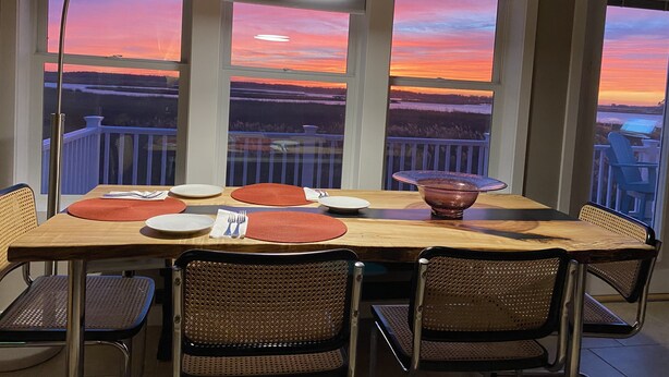 Una mesa de comedor con sillas y vistas a un atardecer a través de grandes ventanales.