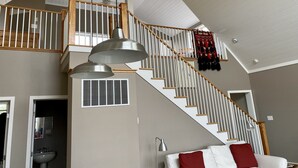 Amplia sala de estar con techo alto, escalera con barandillas de madera y lámpara colgante.