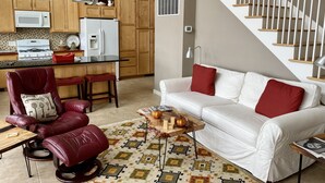 Una cocina moderna con armarios de madera, un refrigerador blanco y un microondas. Hay una silla de cuero rojo y un sofá blanco con cojines rojos. Una mesa de centro de madera con una alfombra estampada está en el centro.