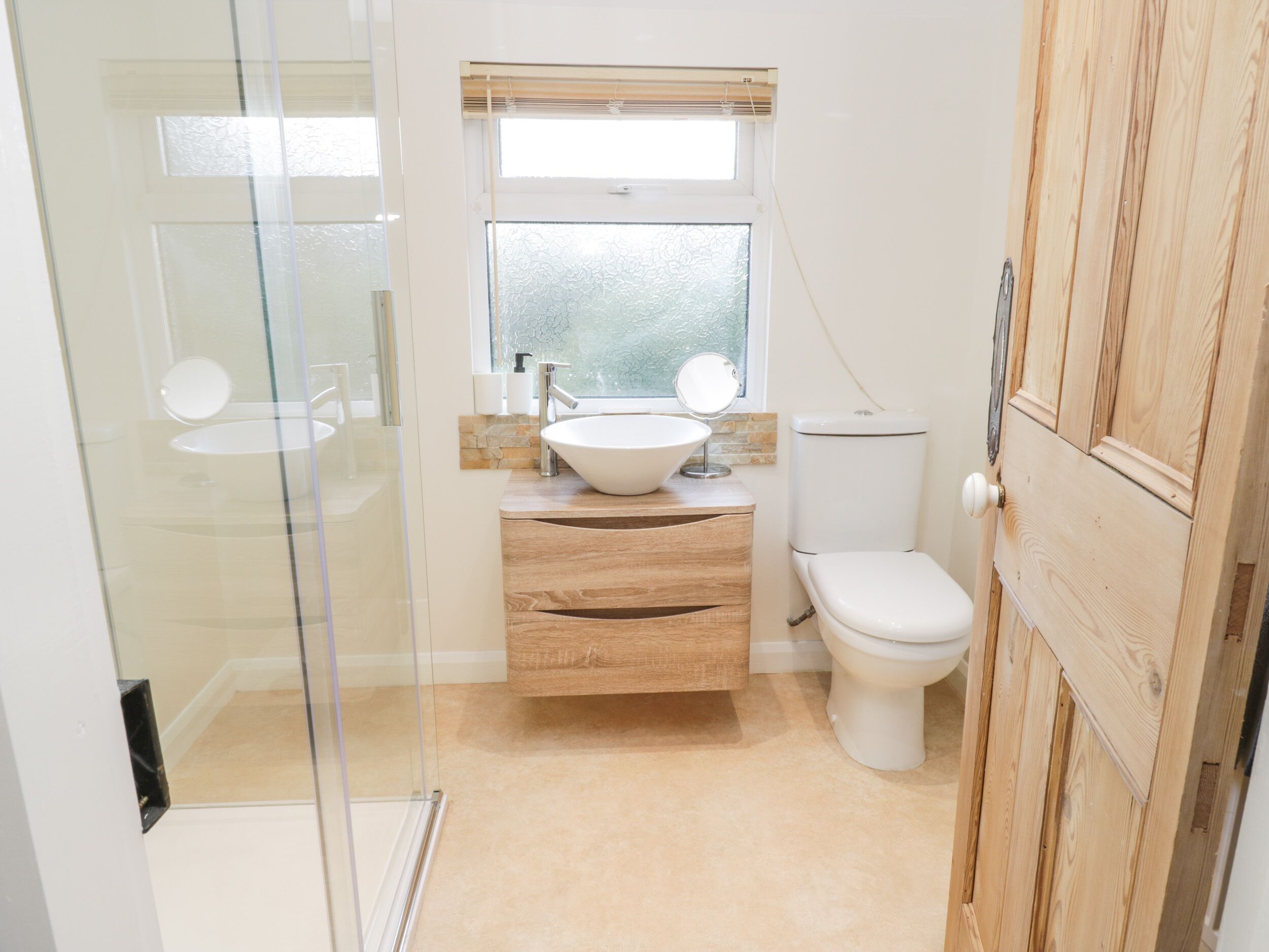A bathroom with a glass shower enclosure, a wooden vanity with a sink, a toilet, and a window above the sink.