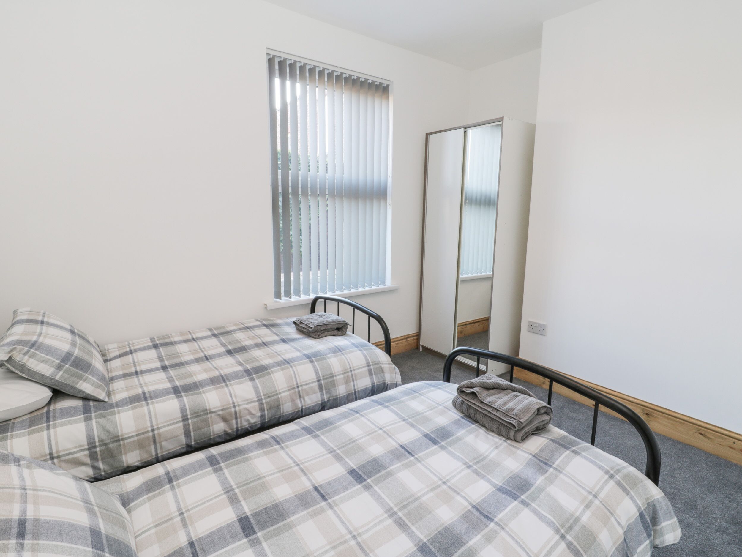 A bedroom with a bed, a window with blinds, a mirror, and a small bedside table.