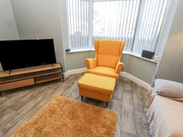 A living room with a flat-screen TV, a yellow armchair, a matching ottoman, a brown rug, and a window with blinds.