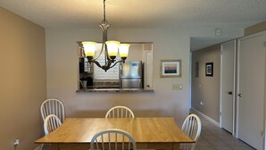 Comedor con mesa de madera y sillas blancas, una lámpara de araña, cocina con refrigerador y una puerta que da a otra habitación.