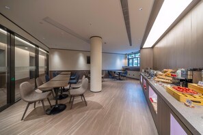 Un salón de conferencias moderno con una amplia mesa y sillas, una zona de cocina con exhibición de alimentos y un gran ventanal que proporciona luz natural.