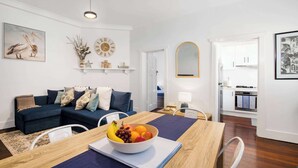 A modern living room with a dining table, a blue sofa, a fruit bowl, and a kitchen in the background.