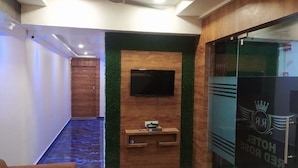 A hotel reception area with a wooden reception desk, a flat-screen TV, and a glass door with the hotel's logo. - Hotel Red Rose (Vadodara)