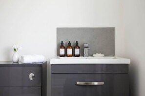 A bathroom vanity with a white countertop, three bottles of toiletries, a white vase with flowers, and a towel. - Stylish 1BR Bolton Apt near Rail & Centre (Bolton)