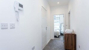 A narrow hallway with a white wall, a door, a bedside table with towels, and a bed in the background.