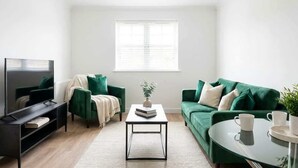 A modern living room with a flat-screen TV, green sofas, a coffee table, and a window with blinds.