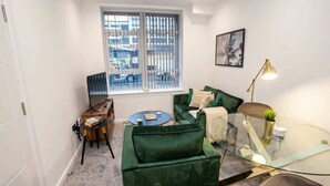 A compact living space with a green sofa, a glass coffee table, a small side table, and a window with blinds.