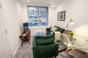 A compact living space with a green sofa, a glass coffee table, a small side table, and a window with blinds. - Stylish 1BR Bolton Apt near Rail & Centre (Bolton)