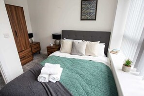 Una cama bien hecha con colcha verde, manta gris y toallas blancas dobladas. A la izquierda, un armario de madera, una mesita de noche con una lámpara y una pequeña planta en maceta en el alféizar.