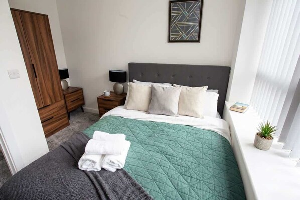 A neatly made bed with a green quilt, a grey blanket, and folded white towels. There is a wooden wardrobe to the left, a bedside table with a lamp, and a small potted plant on the windowsill.
