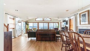 Een ruime woonkamer met een houten piano, een eettafel met stoelen, en grote ramen met uitzicht op de omgeving.