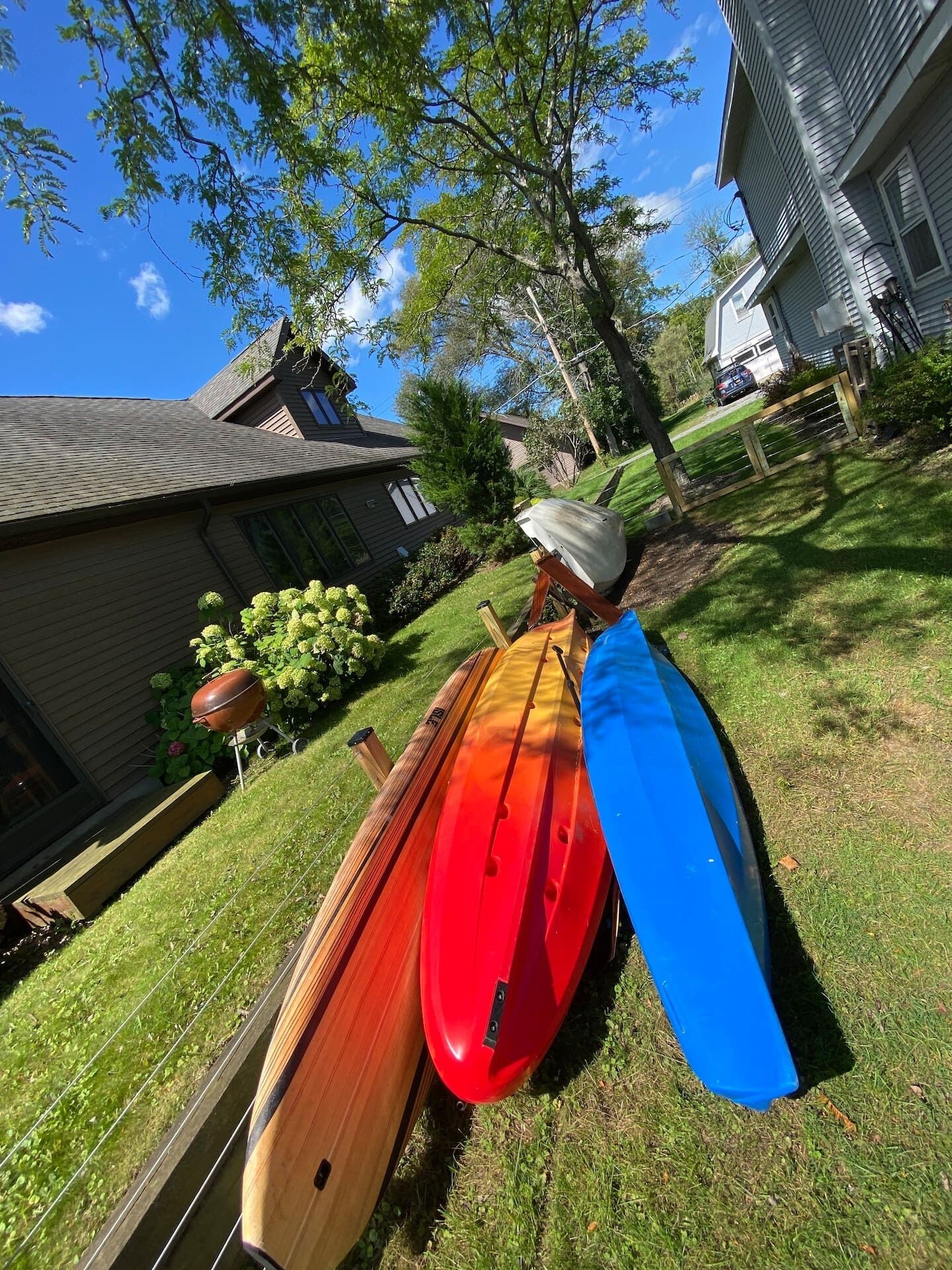 A row of kayaks and a canoe on a grassy lawn with a house in the background.