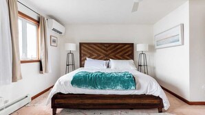 Een slaapkamer met een houten bed, witte beddengoed, een blauwgroene plaid, twee bedlampen, een plafondventilator en een raam met gordijnen.