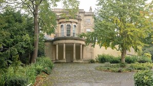 Un bâtiment historique doté d’un portique circulaire central, entouré d’une végétation luxuriante et bordé d’un chemin pavé.
