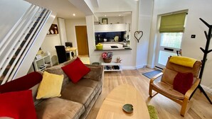 Un salon moderne avec un canapé, un fauteuil et une table basse. On aperçoit une cuisine à l’arrière-plan, avec un coin repas.